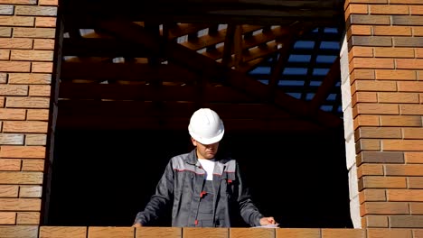 Desde-abajo-la-vista-del-hombre-adulto-en-casco-de-pie-en-el-marco-de-la-ventana-de-la-casa-en-el-sitio-tomando-notas-sobre-papel