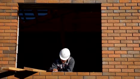 From-below-view-of-building-in-empty-window-frame-of-house-under-construction-standing-in-sunlight-and-taking-notes-on-paper