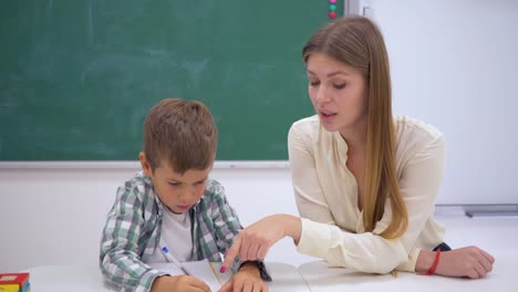 la-escuela-primaria,-el-tutor-profesional-ayuda-a-aprender-a-escribir-al-alumno-en-la-mesa-junto-a-la-Junta