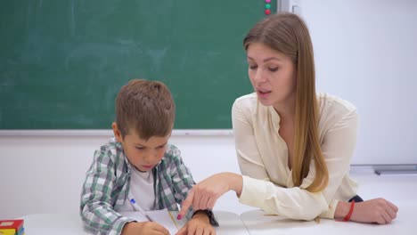 Grundschule,-hilft-professionellen-Tutor-Schulkind-am-Tisch-in-der-Nähe-von-Board-schreiben-lernen