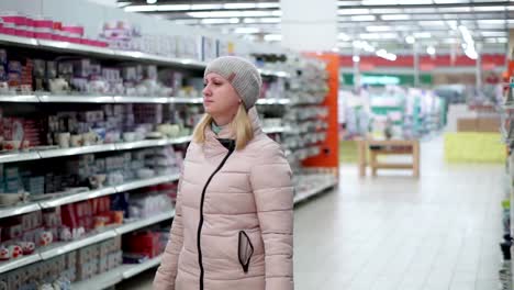 A-woman-in-a-hat-and-a-down-jacket-walks-through-the-supermarket.-She-looks-at-the-goods-on-the-shelves,-chooses-a-purchase.