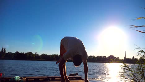 Young-guy-practicing-yoga-moves-and-positions-on-edge-of-wooden-jetty-at-lake-on-sunny-day.-Sporty-man-training-at-nature-with-sunlight-at-background.-Healthy-active-lifestyle.-Slow-motion-Close-up