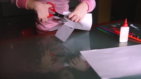 Close-up-shot-of-cute-preschool-girl-in-pink-sweater-cutting-shapes-with-scissors-from-paper,-reflecting-in-glass-table