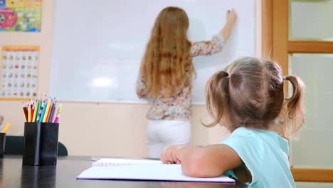 Kleine-süße-Mädchen-sitzt-im-Klassenzimmer-und-Studien-mit-Lehrer-im-Heft