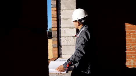 Side-view-of-man-in-hardhat-standing-at-window-in-house-under-construction-looking-at-paper-draft,-4k