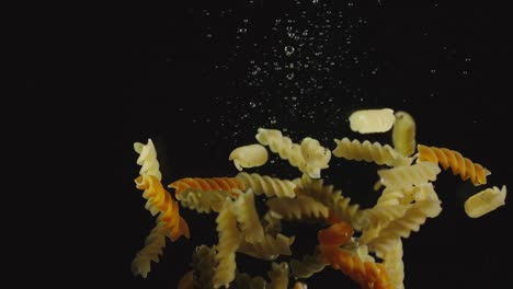 SLOW-MOTION:-Colored-spiral-pasta-fall-under-water-on-black-background