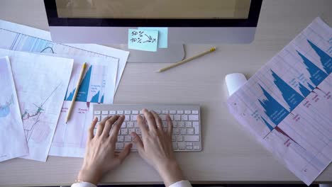 Young-female-employee-is-working,-uses-computer,-sits-at-table,-types-text,-looks-at-monitor,-presses-on-keyboard-with-fingers,-documents-with-graphs-and-diagrams-on-desk-FullHD