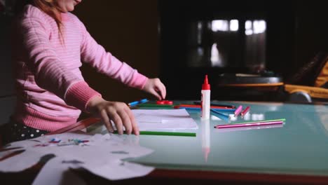 Camera-sliding-left-on-little-cute-Caucasian-girl-child-in-pink-sweater-drawing-with-a-pencil-at-table-with-stationery