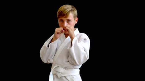 A-young-athlete-in-a-white-kimono-stands-on-a-dark-background.-The-boy-has-blond-hair-and-a-European-look.-Hands-fall,-around-the-waist-there-is-a-white-belt