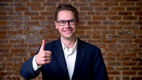 Happy-caucasian-businessman-in-glasses-demonstrating-like-gesture-and-looking-at-camera-straight,-brick-wall-behind