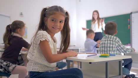 Retrato-de-muchacha-hermosa-estudiante-en-el-escritorio-durante-la-lección-de-educación-en-aula-en-escuela-primaria-de-fondo-desenfocado