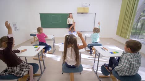 Gruppe-von-Schulkindern-erhöhen-Hände-auf-Lektion-Antworten-beim-Sitzen-am-Schreibtisch-vor-Lehrer-an-Bord-in-der-Grundschule