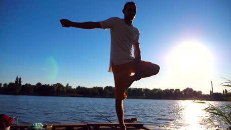 Junger-Kerl-üben-Yogaposition-am-Rande-der-Holzsteg-am-See-am-Sommertag.-Sportlichen-Mann-Übung-in-der-Nähe-von-See-mit-Sonnenlicht-im-Hintergrund.-Gesunden,-aktiven-Lebensstil.-Slow-Motion-hautnah