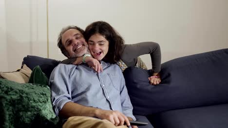 Content-father-sitting-on-couch,-his-daughter-hugging-and-kissing-him