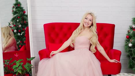 A-blond-woman-in-a-beautiful-dress-and-a-crown-is-posing-in-the-studio.-Red-sofa