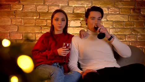 Portrait-of-young-caucasian-couple-sitting-on-sofa-and-drinking-wine-attentively-watching-movie-in-christmas-atmosphere.