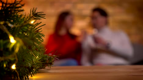 Weihnachtsbaum-mit-hellen-Lichtern-und-unscharfen-junges-Paar-auf-Sofa-sitzen-und-reden-auf-Hintergrund.