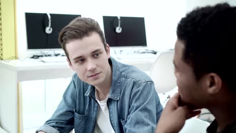 Handsome-Caucasian-student-talking-to-his-African-American-friend-while-sitting-at-desk-in-computer-classroom