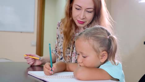 Little-cute-girl-sits-in-classroom-and-studies-with-teacher-in-exercise-book