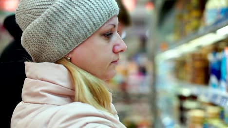 Frau-in-Winterkleidung-in-einem-Supermarkt-Lesen-von-Texten-auf-ein-Glas-von-Lebensmitteln.-Sie-stellt-das-Glas-auf-dem-Regal-des-Kühlschranks.