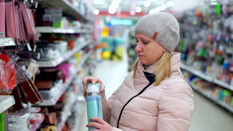 Eine-Frau-mittleren-Alters-in-einen-Hut-und-eine-Daunenjacke-wählt-eine-Thermoskanne-in-einem-Supermarkt.