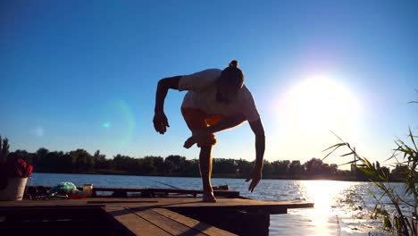 Young-guy-practicing-yoga-moves-and-positions-on-edge-of-wooden-jetty-at-lake-on-sunny-day.-Sporty-man-training-at-nature-with-sunlight-at-background.-Healthy-active-lifestyle.-Slow-motion-Close-up