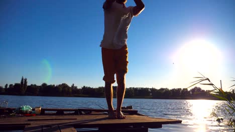 Sporty-man-standing-at-yoga-pose-on-the-edge-of-wooden-jetty-at-lake.-Young-athlete-doing-stretch-exercise-at-nature-on-sunny-summer-day.-Concept-of-healthy-active-lifestyle.-Slow-motion-Close-up