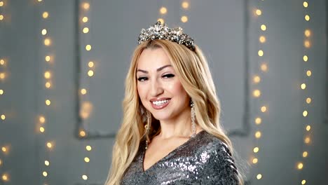 A-blond-woman-in-a-beautiful-dress-and-a-crown-is-posing-in-the-studio.-Red-sofa