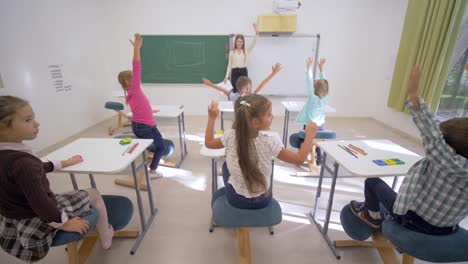 Gruppe-von-Schülern-heben-die-Hand-zur-Beantwortung-an-Lektion-beim-Sitzen-am-Schreibtisch-vor-Erzieher-Blackboard-in-Grundschule