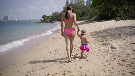 Mama-und-Tochter-gehen-in-rosa-Badeanzügen-am-Meer-entlang.-4K