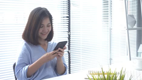 Hermosa-joven-sonriente-mujer-asiática-trabajando-en-ordenador-portátil-mientras-está-sentado-en-una-sala-de-estar-en-casa.-Mujer-de-negocios-asiática-con-teléfono-para-el-trabajo-en-su-oficina-en-casa.-Disfrutando-en-casa.