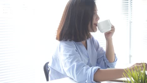 Beautiful-young-smiling-woman-working-on-laptop-while-enjoying-drinking-warm-coffee-sitting-in-a-living-room-at-home.-Enjoying-time-at-home.-Asian-business-woman-working-in-her-home-office.