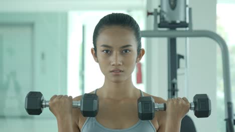 Attractive-shape-young-sporty-focused-fitness-asian-girl-with-ponytail-doing-biceps-exercises-while-sitting-on-the-bench-and-raising-dumbell-in-the-gym