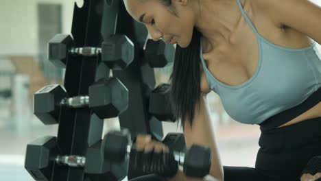 Attractive-shape-young-sporty-focused-fitness-asian-girl-with-ponytail-doing-biceps-exercises-while-sitting-on-the-bench-and-raising-dumbell-in-the-gym