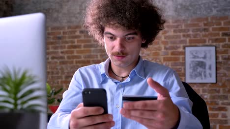 Young-concentrated-man-with-curly-hair-and-mustache-using-credit-card-and-holding-phone-sitting-at-table-in-modern-office