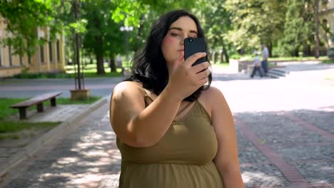 Beautiful-young-overweight-woman-taking-selfie-with-her-phone-and-doing-different-faces-in-camera,-standing-on-street-in-park