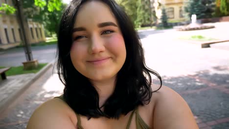 Beautiful-obese-young-woman-recording-video-on-her-web-camera-and-smiling,-standing-on-street-in-park-near-college-building