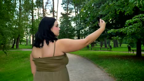 Back-view-of-beautiful-young-obese-woman-taking-selfie-and-walking-in-park-with-trees-and-green-grass,-charming-lady-in-dress