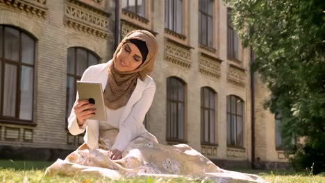 Young-muslim-woman-in-hijab-sitting-on-grass-and-having-video-chat-through-tablet,-showing-location-to-somebody,-building-in-background