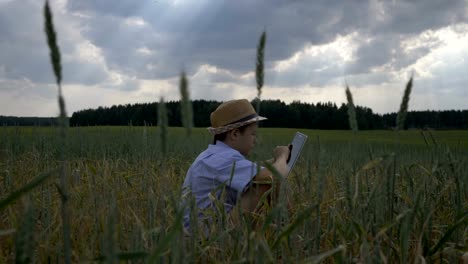 Junge-sitzt-in-einem-Feld-gegen-schöne-Wolken-und-nutzt-eine-Tablette-am-Abend