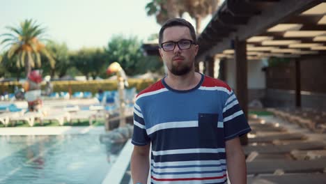 Relaxed-man-is-walking-along-swimming-pool-in-tranquil-yard,-looking-at-camera