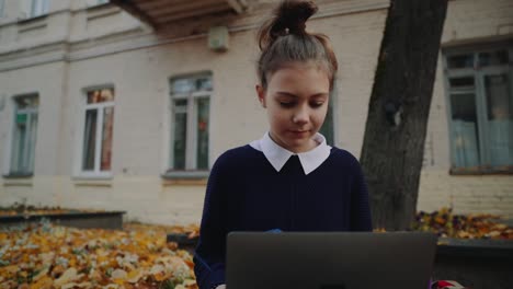 Close-up-pretty-hipster-teen-girl-sitting-on-a-sidewalk-on-autumn-city-street-and-working-laptop-computer.-Schoolgirl-using-notebook-outdoor.-Beautiful-autumn-weather