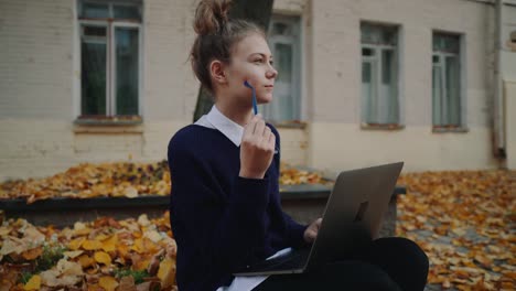 Close-up-pretty-hipster-teen-girl-sitting-on-a-sidewalk-on-autumn-city-street-and-working-laptop-computer.-Schoolgirl-using-notebook-outdoor.-Beautiful-autumn-weather