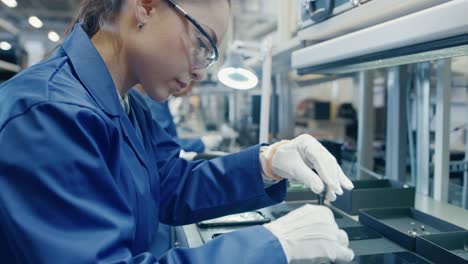 Female-Electronics-Factory-Worker-in-Blue-Work-Coat-and-Protective-Glasses-is-Assembling-Smartphones-with-Screwdriver.-High-Tech-Factory-Facility-with-more-Employees-in-the-Background.