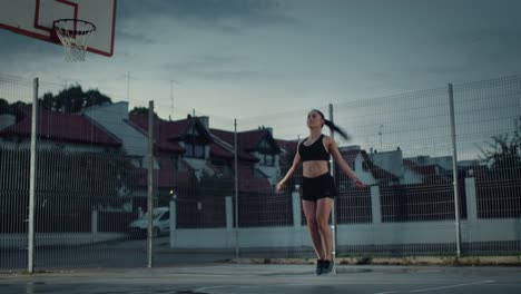 Hermoso-gimnasio-energética-chica-saltando/saltar-cuerda.-Ella-está-haciendo-un-entrenamiento-en-una-cancha-de-baloncesto-al-aire-libre-cercado.-Imágenes-de-noche-después-de-la-lluvia-en-una-zona-de-barrio-residencial.