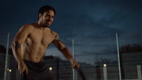 Fuerte-ajuste-camisa-joven-hombre-es-haciendo-ejercicios-con-las-cuerdas-de-la-batalla.-Está-realizando-un-entrenamiento-en-una-cancha-de-baloncesto-al-aire-libre-cercado.-Imágenes-de-noche-después-de-la-lluvia-en-una-zona-de-barrio-residencial.