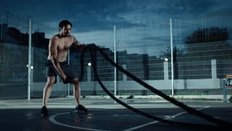 Starke-muskuläre-Fit-Hemd-Jüngling-ist-tun-Übungen-mit-Battle-Ropes.-Er-macht-ein-Training-in-einem-eingezäunten-Basketballfeld-im-Freien.-Abend-Footage-nach-Regen-in-einer-Wohngegend.