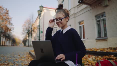 Close-up-pretty-hipster-teen-girl-sitting-on-a-sidewalk-on-autumn-city-street-and-working-laptop-computer.-Schoolgirl-using-notebook-outdoor.-Beautiful-autumn-weather