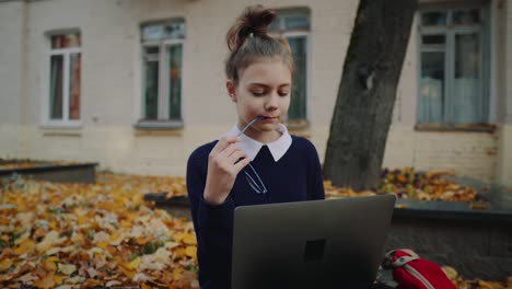 Close-up-pretty-hipster-teen-girl-sitting-on-a-sidewalk-on-autumn-city-street-and-working-laptop-computer.-Schoolgirl-using-notebook-outdoor.-Beautiful-autumn-weather