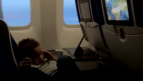 View-of-little-boy-watching-films-in-headset-in-the-aircraft-lying-on-the-seat-against-airplane-window